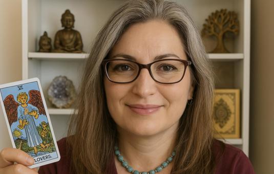 Een vrouw met halflang, grijzend haar en een warme glimlach houdt een tarotkaart omhoog. Ze draagt een bril en een turquoise kralenketting. Op de acht